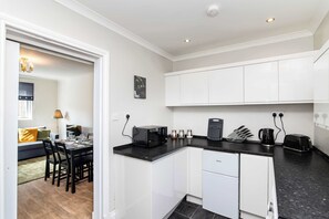 A compact kitchen with white cabinets, a black countertop, a coffee maker, a toaster, and a refrigerator. - 3BR Free Parking Near Hopetoun & Edinburgh Airport (Bo'Ness)