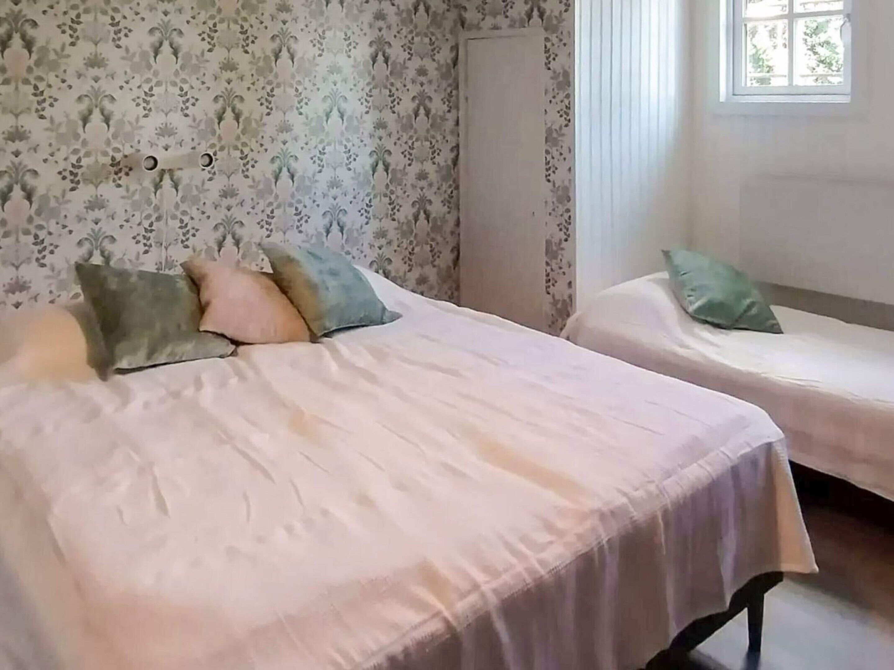 A room with two beds, one with a white bedspread and the other with a light-colored bedspread. There are pillows on the beds, and a window with curtains is visible.
