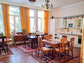 Un coin repas avec une table et des chaises en bois, une cuisine avec des meubles blancs et de grandes fenĂȘtres dotĂ©es de rideaux.