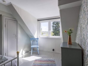 Une pièce avec un plafond en pente, une fenêtre donnant sur les arbres, une chaise, un lit et une petite table sur laquelle est posé un vase.
