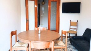 Une pièce comprenant une table ronde en bois, quatre chaises, un canapé noir, un téléviseur fixé au mur et une porte dotée d’un panneau en verre dépoli.
