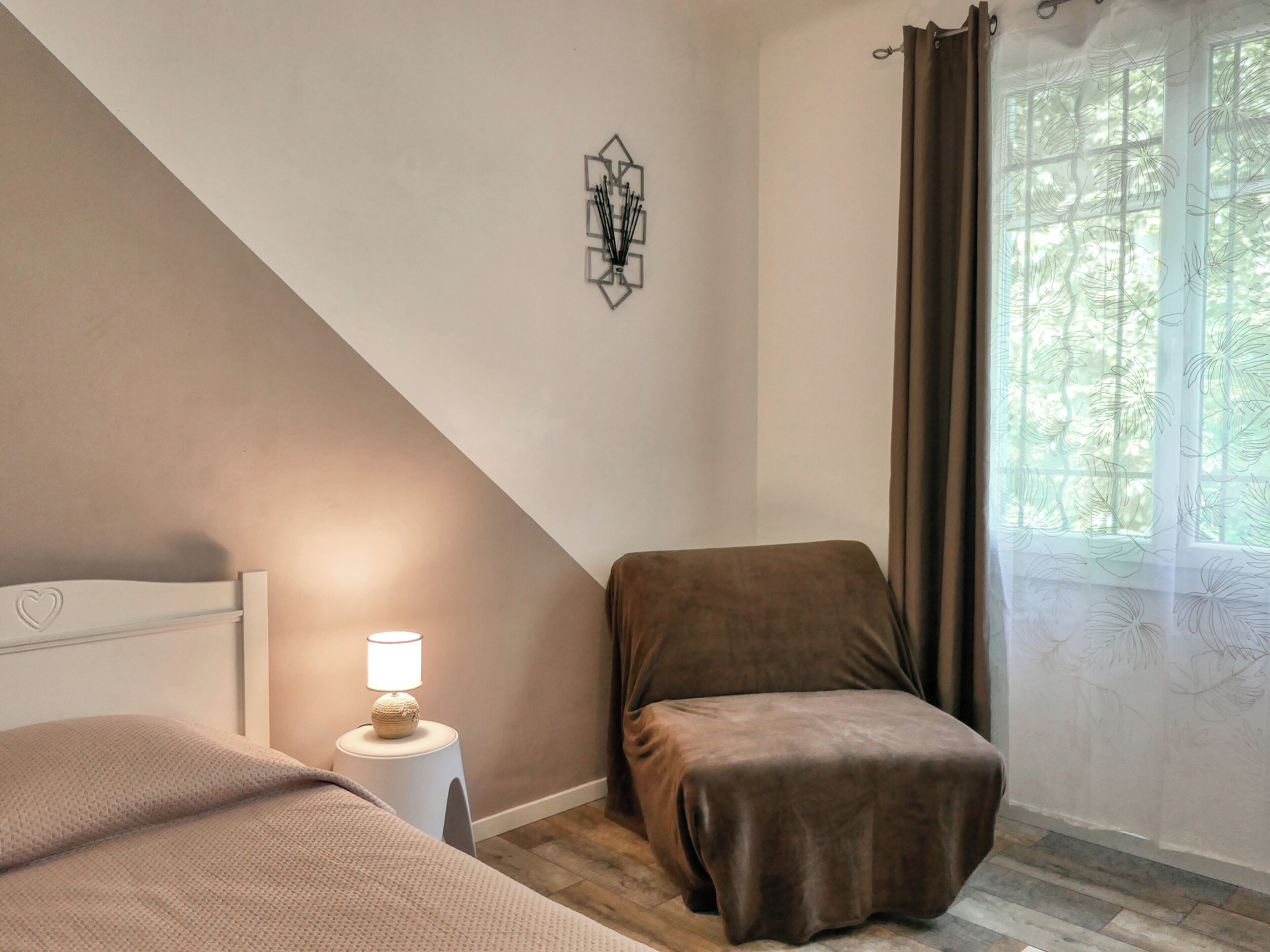 Une chambre à coucher avec un lit, une table de chevet avec une lampe, une fenêtre avec des rideaux et un tableau accroché au mur.