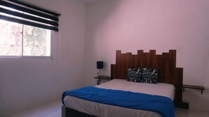 Une chambre à coucher comprenant un lit, une table de chevet, une lampe, une fenêtre avec des stores et une tête de lit en bois.