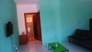 Une pièce avec un mur vert, une porte en bois donnant sur une salle de bain, une table verte sur laquelle se trouve un ananas, un canapé et un téléviseur fixé au mur.