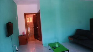 A room with a green wall, a wooden door leading to a bathroom, a green table with a pineapple, a couch, and a TV on the wall. - Downstairs Oasis/Private Pool/ High-Speed WiFi (Punta Cana)