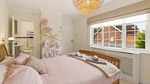 Ein Schlafzimmer mit Bett, Fenster mit Blick auf ein Backsteinhaus, Heizkörper und Wand mit tropischem Pflanzenwandbild.