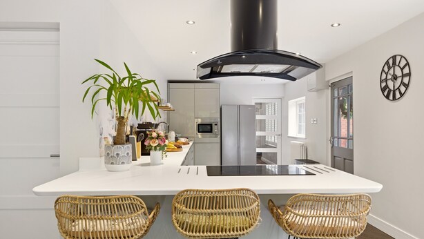 A modern kitchen with a central island, three wicker chairs, a potted plant, and a clock on the wall. - New! Central 3BR near Kenilworth Castle and A46 (Kenilworth)