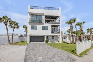 A modern two-story house with a garage, a balcony, and palm trees in the yard. - The White House Pool Oceanviews! Rooftop Deck (New Smyrna Beach)