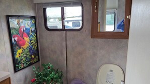 A compact bathroom with a toilet, a small step stool, a framed painting, a window, and a mirror.