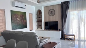 A modern living room with a sofa, TV, air conditioning unit, artwork, shelves, and a window with curtains. - Luxury 3-bedroom Pool Villa in peaceful Hua Hin (Hua Hin)