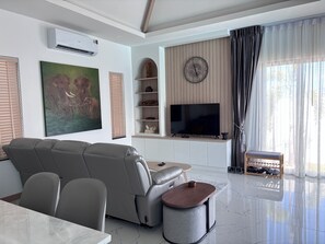 A modern living room with a sofa, TV, air conditioning unit, artwork, shelves, and a window with curtains. - Luxury 3-bedroom Pool Villa in peaceful Hua Hin (Hua Hin)
