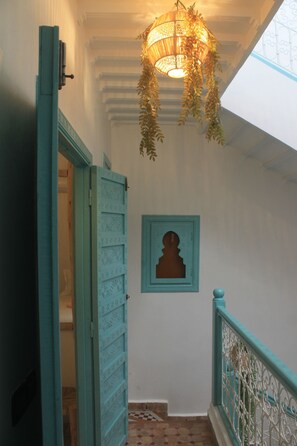 Un couloir avec une porte de couleur turquoise, un luminaire suspendu et une silhouette encadrée sur le mur.