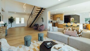 A spacious living area with a staircase, a sofa, ottomans, a coffee table, and a kitchen in the background. - The Superstar | Waterfront Pool & Pickleball (Hot Springs National Park)