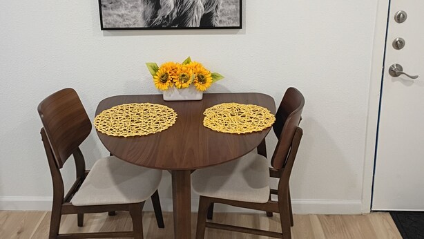 Een eetgedeelte met een ronde tafel, twee stoelen, een lijst met een dierenafbeelding en een vaas met zonnebloemen.