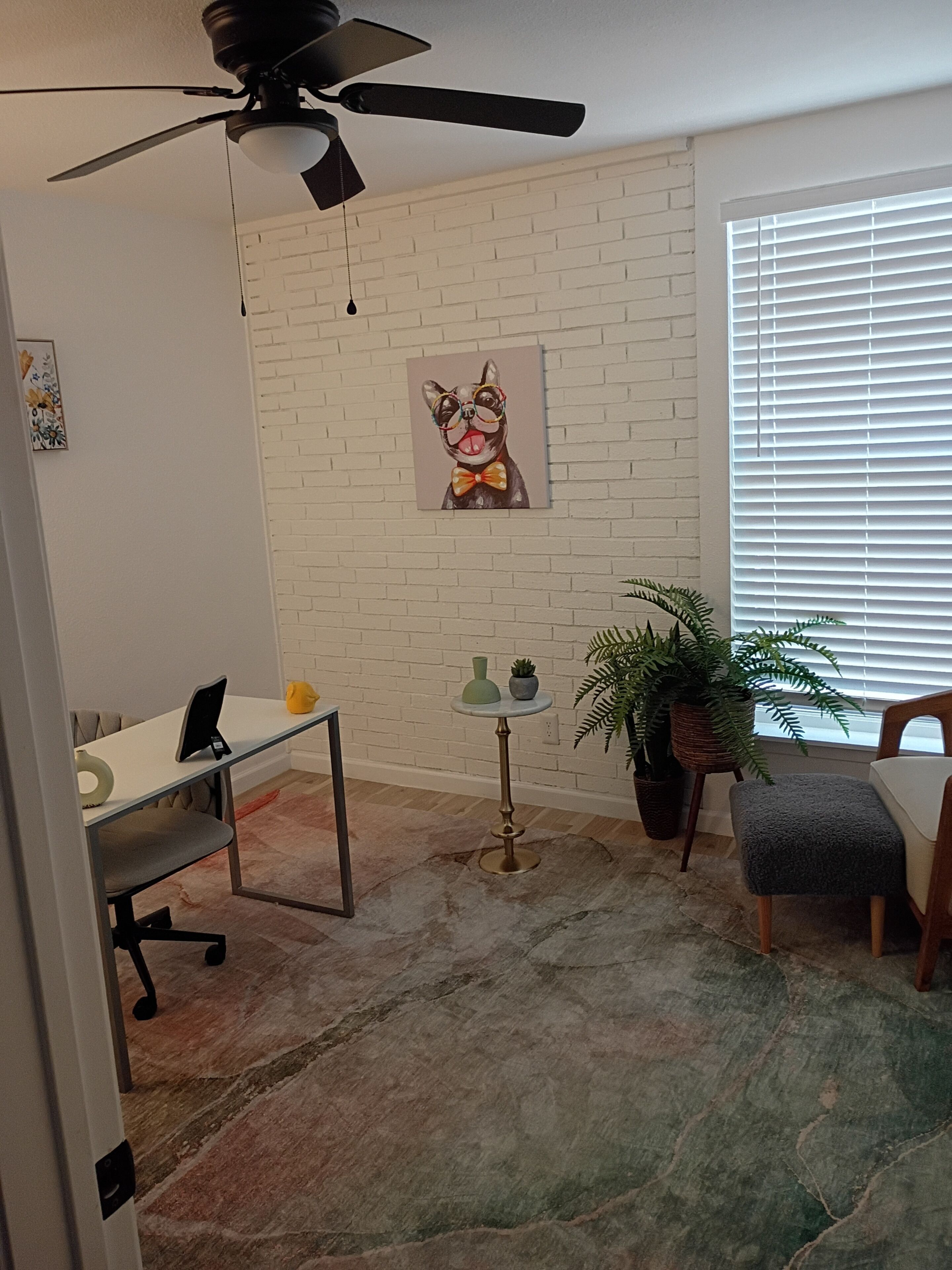 A room with a white brick wall, a ceiling fan, a painting of a dog, a small table with a vase, a potted plant, and a window with blinds.