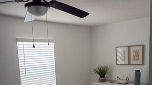 Een slaapkamer met een bed, een dressoir, een plafondventilator, een raam met jaloezieën en twee foto's in lijsten aan de muur.