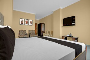 Quarto de hotel com uma cama grande, duas poltronas estampadas, uma televisão e uma mesinha com chaleira.