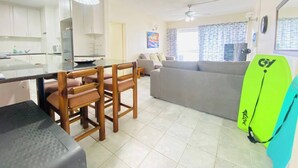 Eine moderne Küche mit weißen Schränken, ein Essbereich mit Holzsesseln, ein graues Sofa und ein Deckenventilator.
