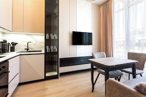 A modern kitchen with a built-in oven, a dining area with a table and chairs, a flat-screen TV mounted on the wall, and a large window with curtains.