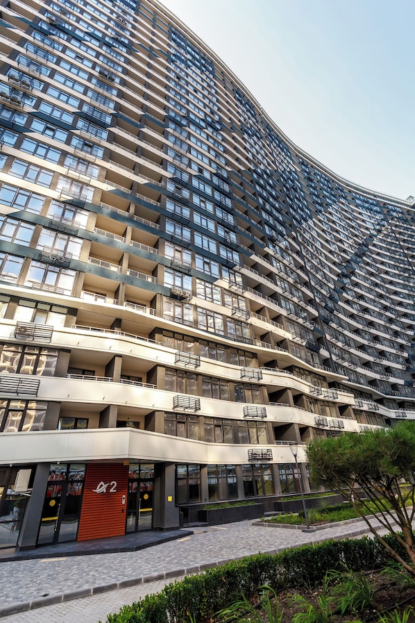 A high-rise building with multiple balconies, a prominent entrance with a red door, and a landscaped area in front.