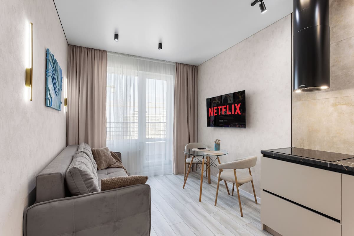 A modern living room with a sofa, a dining table with chairs, a kitchen area, and a wall-mounted TV displaying 'NETFLIX'.