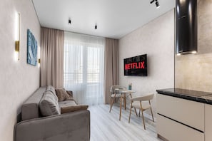 A modern living room with a sofa, a dining table with chairs, a kitchen area, and a wall-mounted TV displaying 'NETFLIX'.