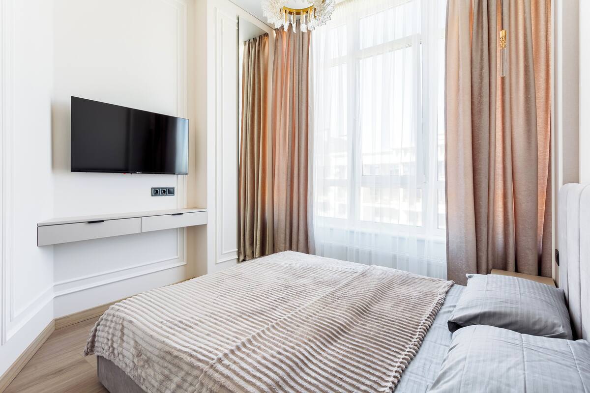 A modern bedroom with a flat-screen TV, a bed with a striped blanket, and sheer curtains.