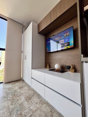Ein modernes Wohnzimmer mit einem an einer Holzwand montierten Flachbildfernseher, einer weißen Unterhaltungseinheit darunter und einem großen Fenster, das natürliches Licht hereinlässt.