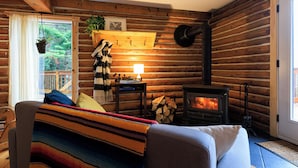 A log cabin interior with a sofa, a fireplace, a hanging plant, and a window with curtains. - Log cabin Laurentians | 5 bedrooms | trails, fireplace, spa, pets allowed (Sainte-Lucie-des-Laurentides)