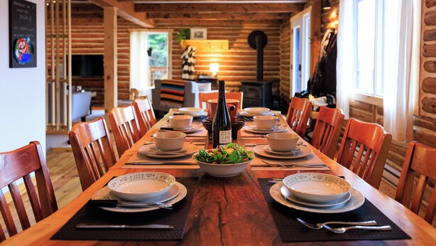A wooden dining table set with plates, bowls, and a bottle of wine, surrounded by chairs in a log cabin interior. - Log cabin Laurentians | 5 bedrooms | trails, fireplace, spa, pets allowed (Sainte-Lucie-des-Laurentides)