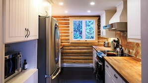 A compact kitchen with a refrigerator, stove, and microwave. A window with a view of trees is visible. - Log cabin Laurentians | 5 bedrooms | trails, fireplace, spa, pets allowed (Sainte-Lucie-des-Laurentides)