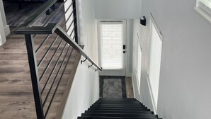 Een trap met een metalen leuning die leidt naar een plateau, een bed met een blauwe sprei, en een raam met jaloezieën.