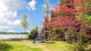 Une aire de jeux avec un toboggan et des balançoires, entourée d’arbres et bordée d’un lac.