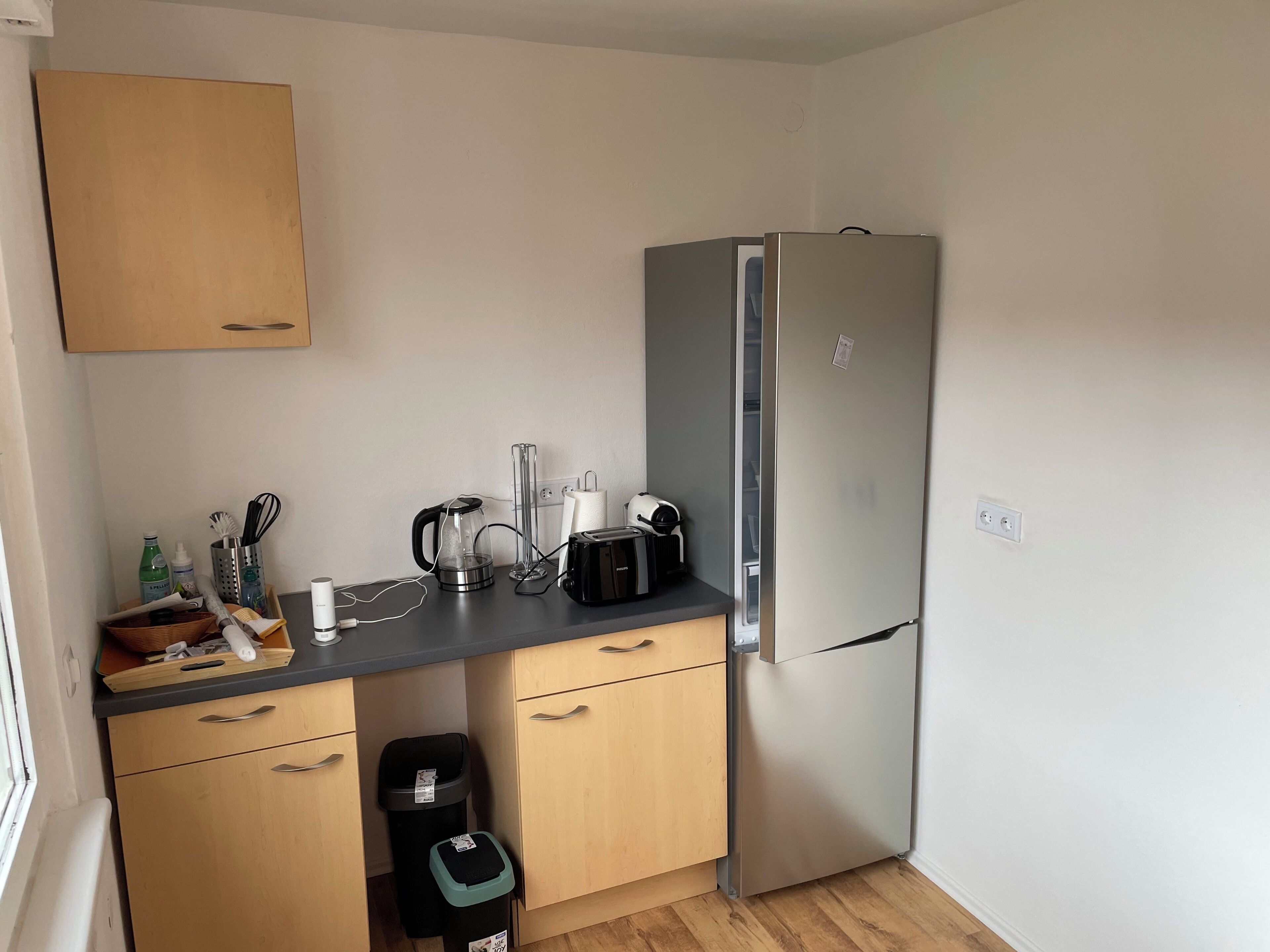 Een kleine keuken met een koelkast, waterkoker, broodrooster en een prullenbak.
