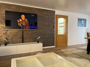 A modern living room with a flat-screen TV mounted on a textured wall, a wooden door, and a small table with a chair. - Vacation apartment in the beautiful Lahn Valley near Bad Ems/Koblenz - 2 identical double rooms (Fachbach)
