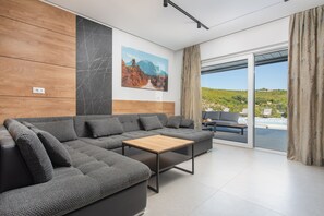 A modern living room with a sectional sofa, a wooden coffee table, and a large window offering a view of a pool and greenery. - Luxury villa Natali with pool, terrace and sea view (Podstrana - Sita)