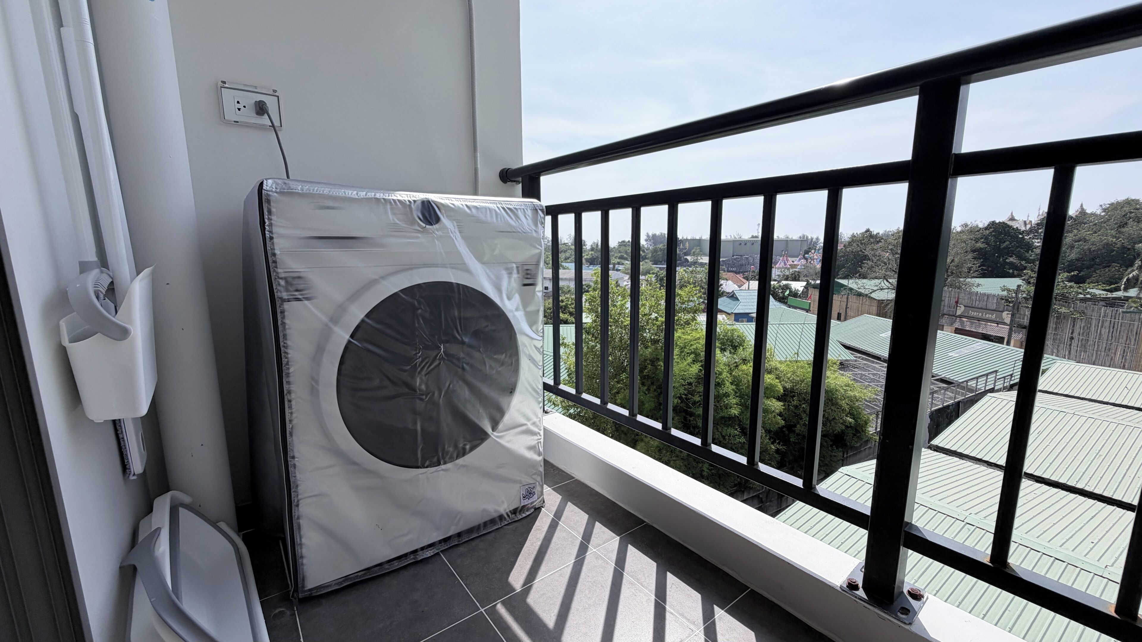 Een balkon met een grote airconditioning, een stopcontact en uitzicht op een park met gebouwen.