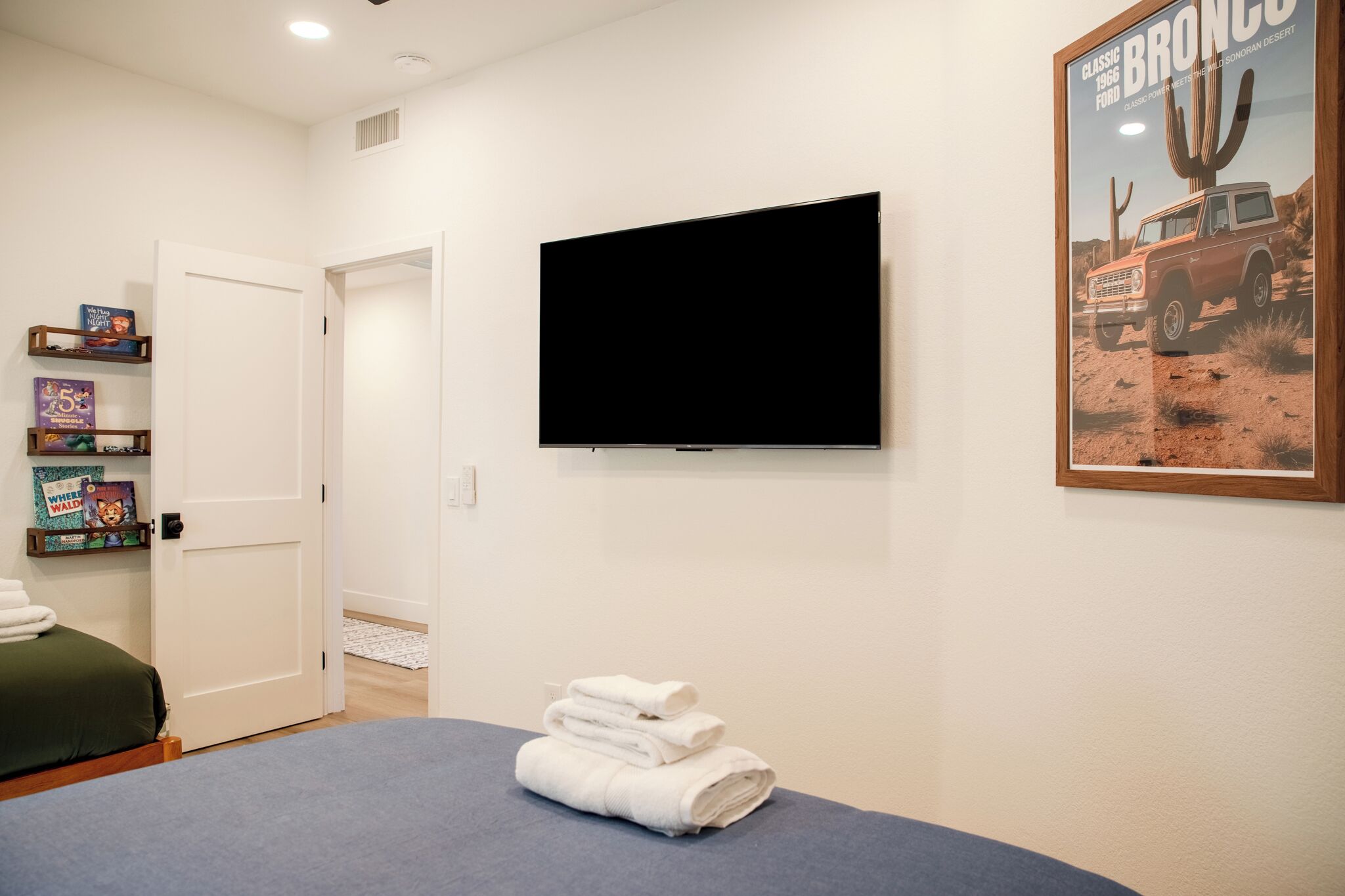 Ein Hotelzimmer mit Bett, einem an der Wand montierten Flachbildfernseher, einer Tür und einem Poster eines klassischen Ford Bronco.