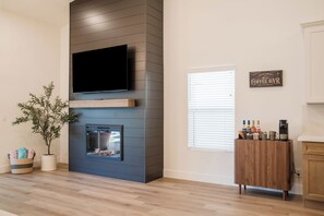 Ein modernes Wohnzimmer mit einem flachbildfernseher an einer dunklen Wand, einem Kamin, einer Kaffeebar mit diversen Utensilien und einer Topfpflanze.