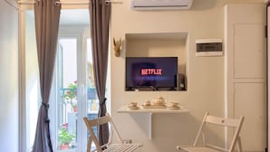 Quarto com televisão exibindo 'NETFLIX', uma mesa pequena com um bule, duas cadeiras e uma varanda com plantas.