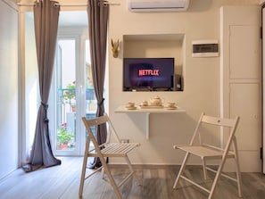 Ein Zimmer mit Fernseher, der 'NETFLIX' anzeigt, einem kleinen Tisch mit Teekanne, zwei Stühlen und einem Balkon mit Pflanzen.