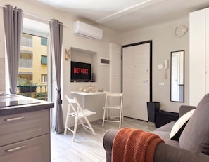 Ein modernes Apartment mit Küchenzeile, einer Essecke mit kleinem Tisch und Stühlen, einem Wohnzimmer mit Sofa, einem an der Wand montierten Fernseher und einer Tür mit Telefon und Spiegel.
