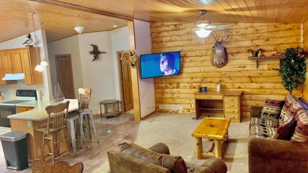 Un accogliente interno di una baita con zona pranzo, soggiorno con divano, televisione, pareti in legno e ventilatore a soffitto.