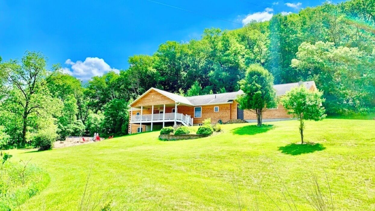 A two-story wooden house with a porch, surrounded by a grassy field and dense trees.