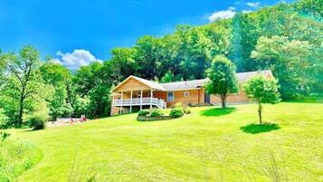 A two-story wooden house with a porch, surrounded by a grassy field and dense trees.