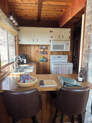 A wooden cabin-style kitchen with a wooden ceiling, white cabinets, a microwave, a stove, a sink, and a dining area with two chairs.