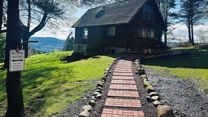 A wooden house with a brick pathway leading to it, surrounded by trees and a grassy area.