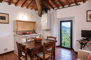 Un coin repas avec une table en bois, des chaises, un panier de fruits, une cuisine avec un fourneau blanc et un balcon avec vue.