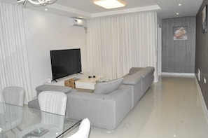 A modern living room with a grey sofa, a glass coffee table, a flat-screen TV, and a dining area with a glass table and chairs.