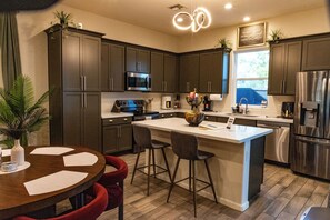 A modern kitchen with dark cabinets, stainless steel appliances, a central island with bar stools, and a dining table with red chairs. - Unbelievable Oasis with Private Pool, Spacious and Resort Home (Goodyear)